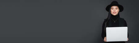 Asian model in fedora hat holding laptop and looking at camera isolated on dark grey, bannerの写真素材