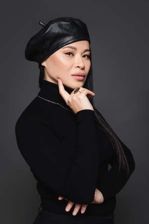 Stylish asian model in beret looking at camera isolated on dark grayの写真素材