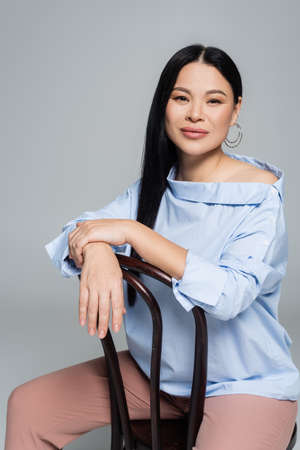 Happy asian woman in blouse sitting on chair isolated on grayの写真素材