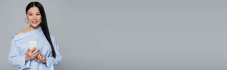 Stylish asian woman in blouse using cellphone and looking at camera isolated on grey, bannerの写真素材