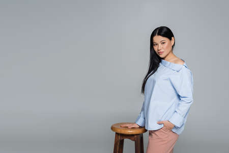 Asian model in blue blouse posing near chair isolated on grayの写真素材
