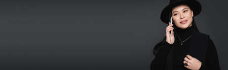 Stylish asian woman in fedora hat and jacket talking on smartphone isolated on dark grey, bannerの写真素材