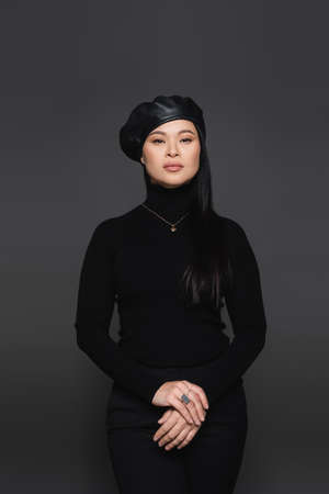 Fashionable asian woman in beret posing and looking at camera isolated on dark grayの写真素材