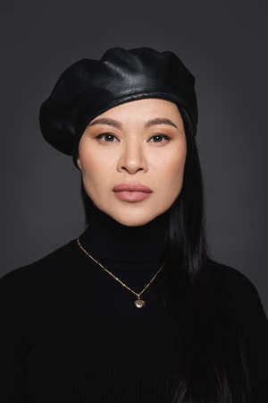 Asian model in beret and turtleneck looking at camera isolated on dark grayの写真素材