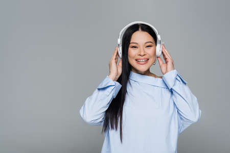 Cheerful asian woman in blouse listening music in headphones isolated on grayの写真素材