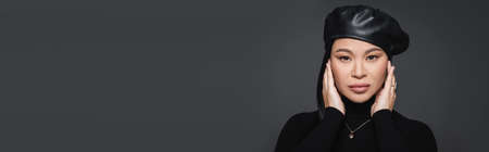 Asian model in beret touching face and looking at camera isolated on dark grey, bannerの写真素材