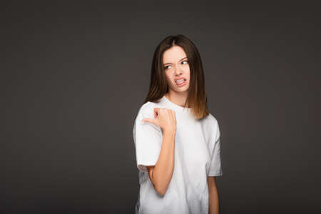 young woman feeling disgusted, sticking out tongue and pointing with thumb isolated on dark gray