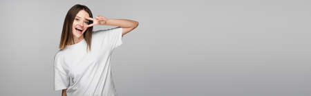 cheerful woman in white t-shirt showing victory sign near face isolated on grey, bannerの写真素材