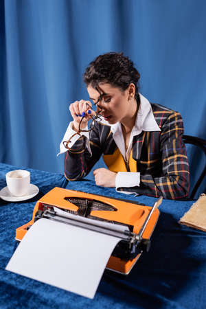 tired journalist sitting with closed eyes near coffee cup and vintage typewriter on blue backgroundの写真素材