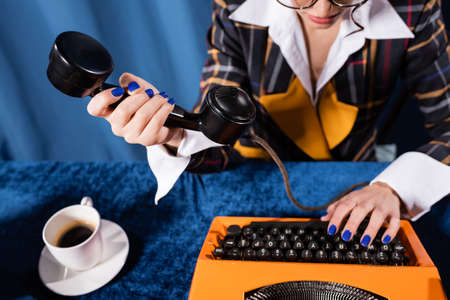 partial view of stylish newswoman holding handset while typing on vintage typewriter on blue backgroundの写真素材