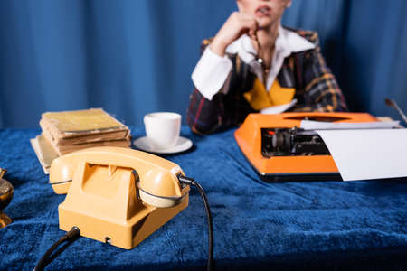 partial view of blurred newswoman near vintage telephone, typewriter and books on blue velour tableclothの写真素材