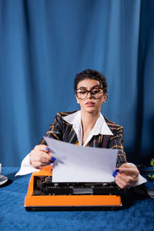 journalist in vintage style clothes holding empty paper near typewriter on blue backgroundの写真素材
