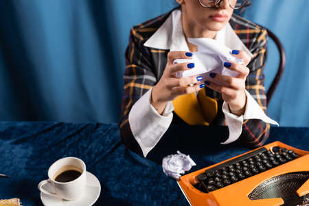 partial view of stylish woman with crumpled paper near vintage typewriter and and coffee cup on blue backgroundの写真素材