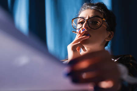 woman in vintage eyeglasses looking at blurred paper while smoking on blue backgroundの写真素材