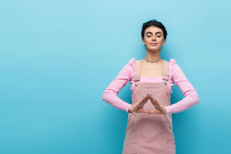positive woman in pastel clothes relaxing in meditation pose isolated on blueの写真素材