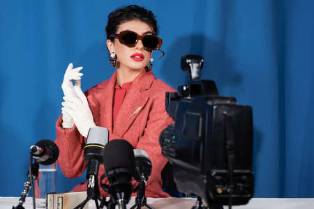 blurred digital camera and microphones near retro style woman giving interview on blue background