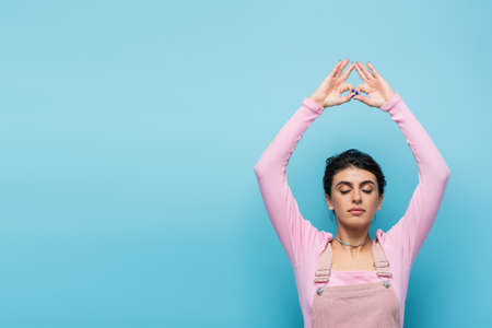 young woman with closed eyes meditating with raised hands and jnana mudra gesture isolated on blueの写真素材