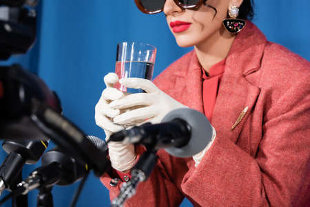 partial view of retro style woman in white gloves holding glass of water near blurred microphones on blue backgroundの写真素材