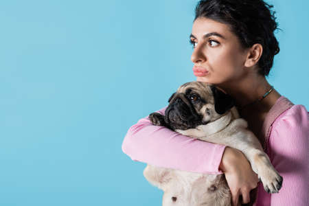 thoughtful brunette woman embracing pug dog while looking away isolated on blueの写真素材