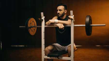 Bearded sportsman sitting on flat bench near barbell on dark backgroundの写真素材
