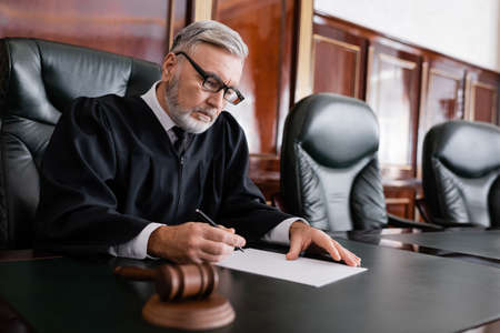 senior judge in robe and eyeglasses holding pen near paper and blurred gavelの写真素材