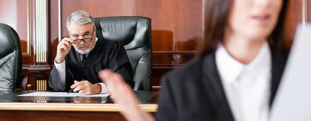 senior judge adjusting eyeglasses while looking at prosecutor on blurred foreground, banner
