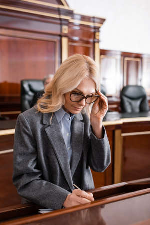 blonde advocate adjusting eyeglasses while writing in courtroom