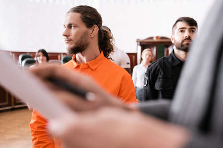 accused man looking away near guard and blurred prosecutor in courtroom
