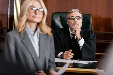 grey-haired judge in robe and eyeglasses near blurred advocate in courtroom