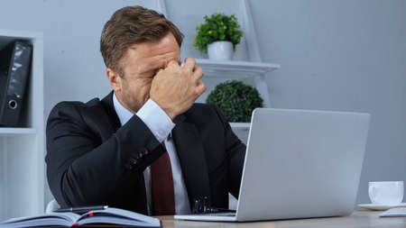 exhausted businessman rubbing eyes while suffering from migraine near laptop