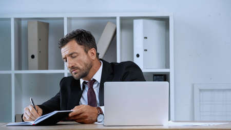 businessman working near laptop and writing in notebook in office