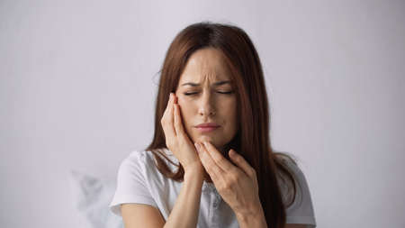 unhappy woman with toothache touching cheek and frowning with closed eyes on grey backgroundの写真素材