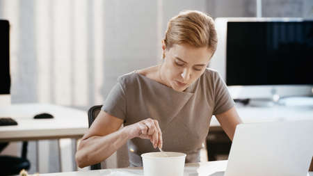 Businesswoman holding fork near takeaway lunch and laptop in office