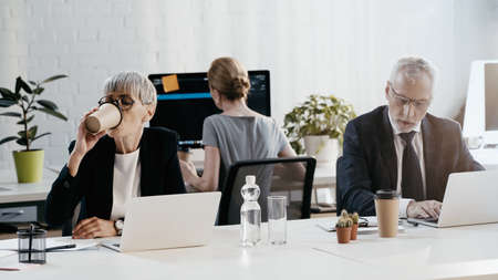 Businesswoman drinking coffee near laptop and documents in officeの写真素材