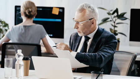 Businessman holding documents and pen near laptop in officeの写真素材