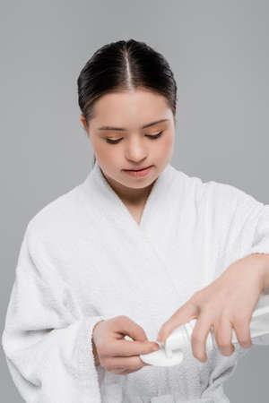 Young woman with down syndrome in bathrobe pouring cleansing foam on cotton pad isolated on greyの写真素材