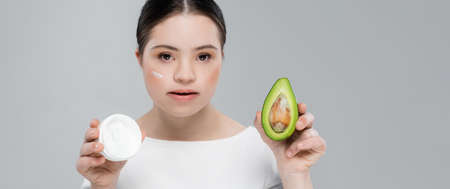 Woman with down syndrome and cream on face holding avocado isolated on grey, banner