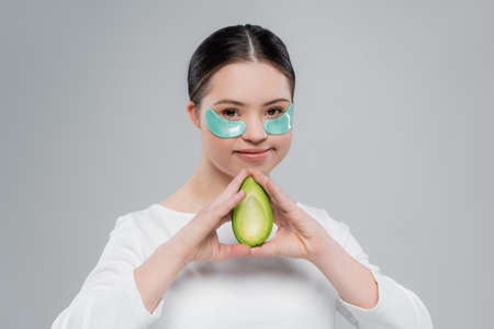 Smiling woman with down syndrome and eye patches holding avocado isolated on greyの写真素材