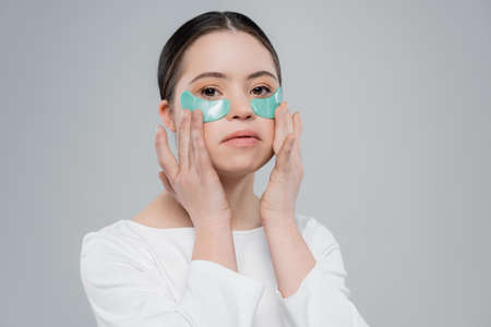 Young woman with down syndrome applying eye patches and looking at camera isolated on grey