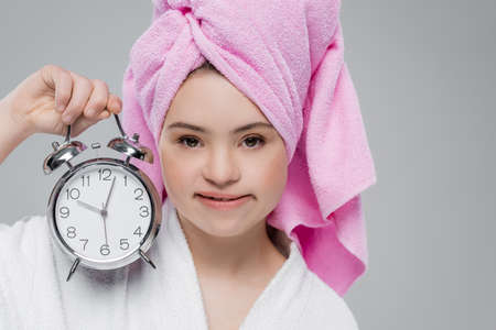 Woman with down syndrome in bathrobe and towel holding alarm clock isolated on greyの写真素材