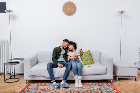 pregnant african american woman sitting with happy husband on couch in modern apartmentの写真素材