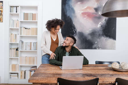 happy pregnant african american woman hugging husband working remotely on laptop