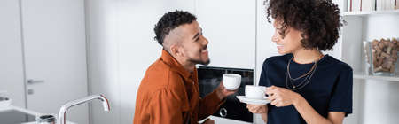 happy african american couple holding cups of coffee in kitchen, banner