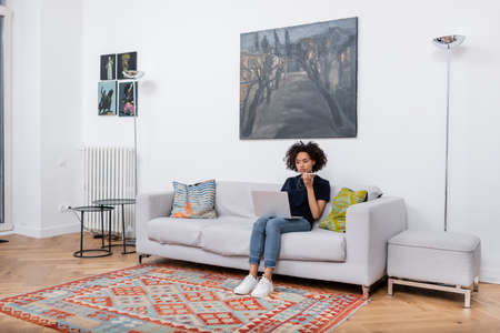 young african american woman sitting on couch and recording voice message on mobile phone while...