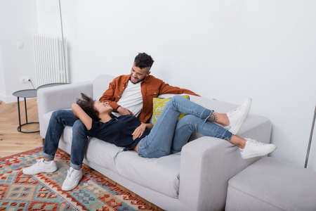 cheerful young african american couple looking at each other while resting on couch in modern...