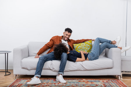 happy african american man looking at girlfriend resting on couch in modern apartmentの写真素材