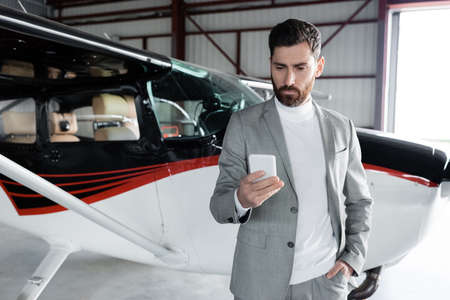 bearded man in formal wear using smartphone and standing with hand in pocket near modern helicopter
