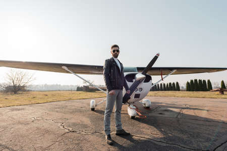full length of bearded pilot in stylish sunglasses and leather jacket standing near modern aircraft