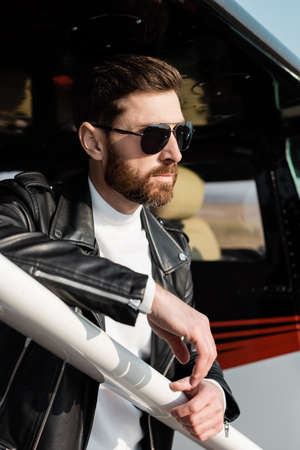 confident bearded man in sunglasses and black leather jacket near helicopter