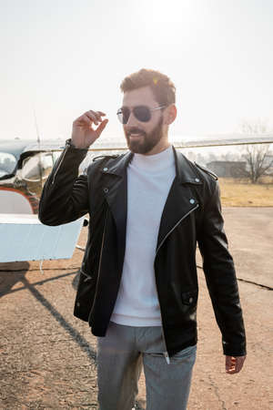 positive pilot in leather jacket and stylish sunglasses standing near helicopterの写真素材